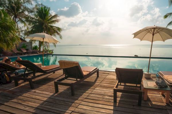 A tranquil beach scene with lounge chairs, umbrellas, and a wooden deck overlooking a pool and ocean under a cloudy sky.