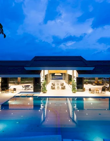 The image shows a modern building with an open design, featuring a large illuminated swimming pool and outdoor seating under a twilight sky.