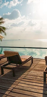 A serene poolside scene with lounge chairs, parasols, and palm trees overlooking a calm ocean under a partly cloudy sky.