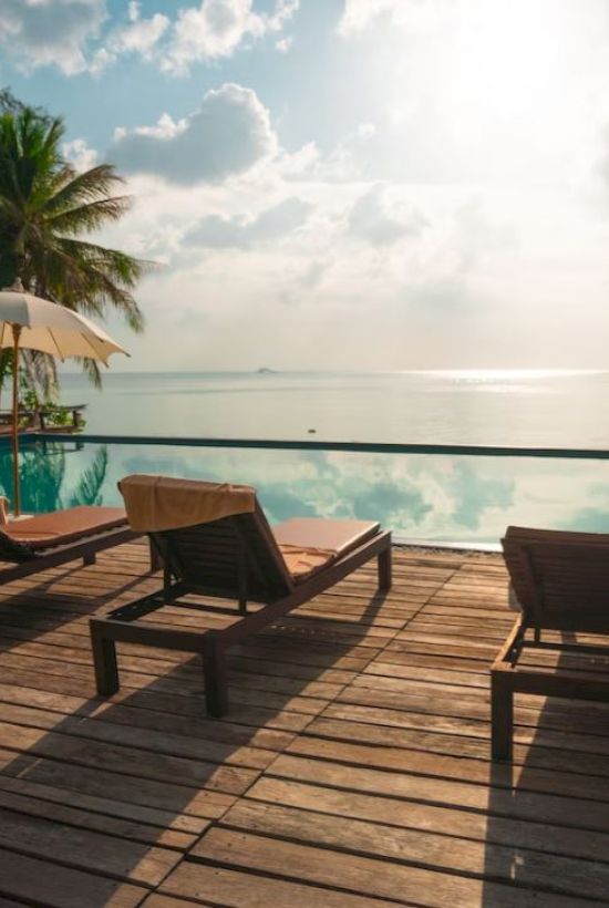 A serene poolside scene with lounge chairs, umbrellas, and palm trees overlooking a calm body of water under a partly cloudy sky.