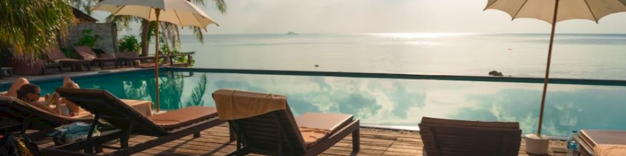 Lounge chairs and umbrellas by a serene pool with a beautiful ocean view, surrounded by palm trees under a partly cloudy sky.
