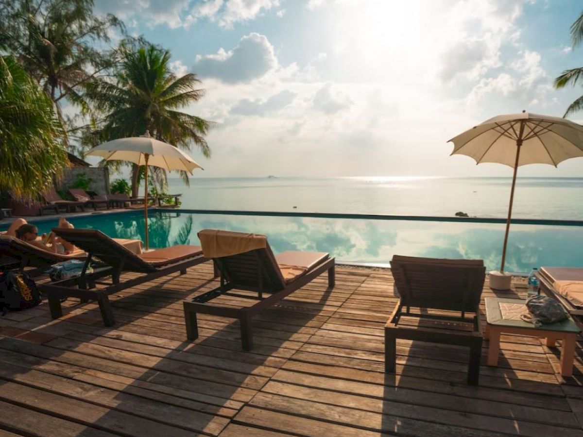 A serene poolside area with lounge chairs, umbrellas, and palm trees overlooking a calm ocean under a partly cloudy sky.