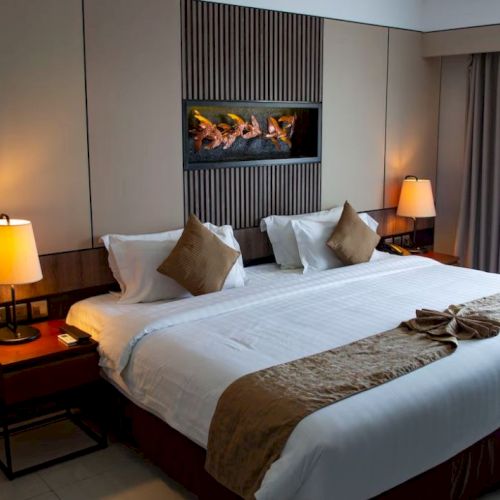 A cozy hotel room with a large bed, decorative pillows, bedside lamps, and a wall-mounted fireplace.
