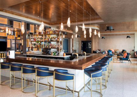 The image shows a modern bar with blue stools, overhead lights, and a fully stocked back bar, set in a spacious, stylish interior.