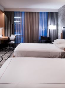 The image shows a modern hotel room with two beds, a wall-mounted TV, a desk with a chair, and stylish lighting and decor.