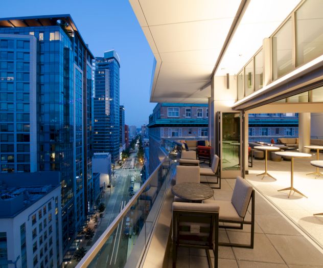 A modern rooftop terrace overlooks a city street with tall buildings, featuring outdoor seating and ambient lighting at dusk.