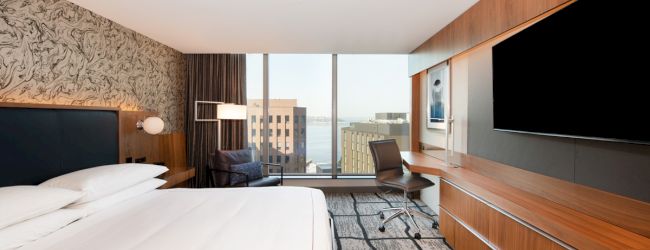 The image shows a modern hotel room with a large bed, desk, TV, and a window view of city buildings.
