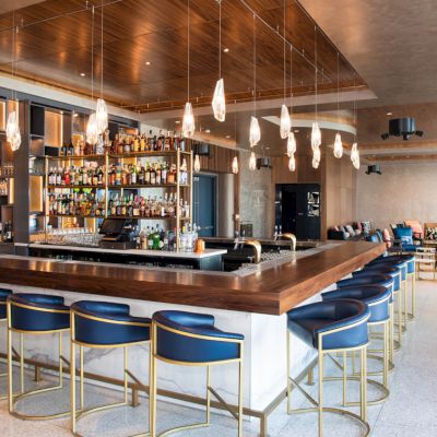 The image features a modern bar with a stocked bottle shelf, elegant lighting, blue bar stools, and a comfortable seating area in a stylish setting.