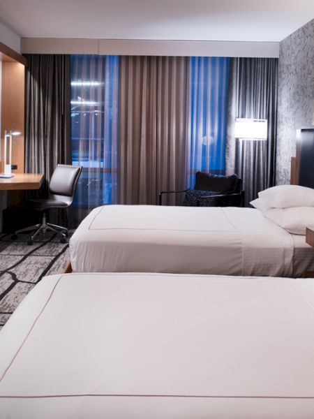 A hotel room with two beds, a large TV on the wall, and a desk with a chair. It's well-lit with modern decor and a patterned carpet.