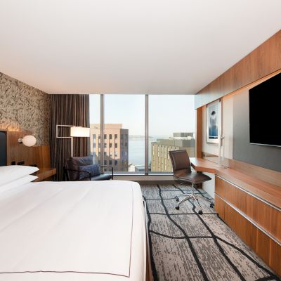 A modern hotel room features a large bed, wall-mounted TV, desk, and city view through a large window.
