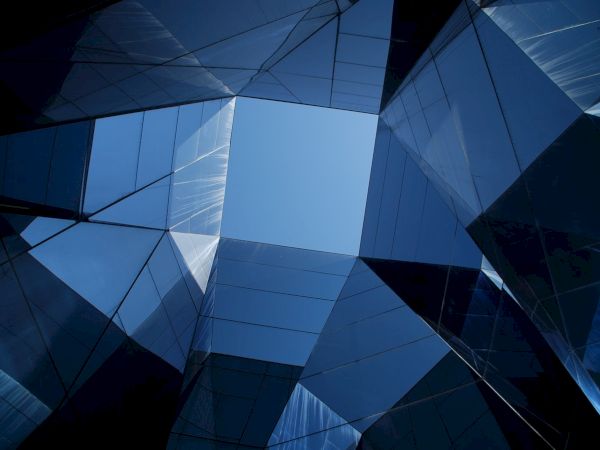 The image shows a view of glass skyscrapers from below, creating a geometric pattern with a clear blue sky at the center.