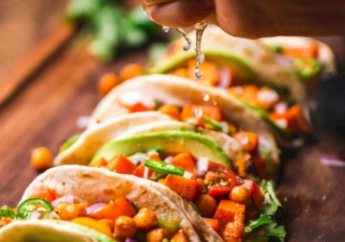 A person is squeezing lime over a row of chickpea and vegetable tacos on a wooden board, garnished with avocado and cilantro.