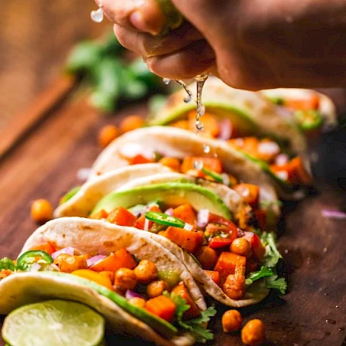 A person is squeezing lime over a row of chickpea and vegetable tacos on a wooden board, garnished with avocado and cilantro.