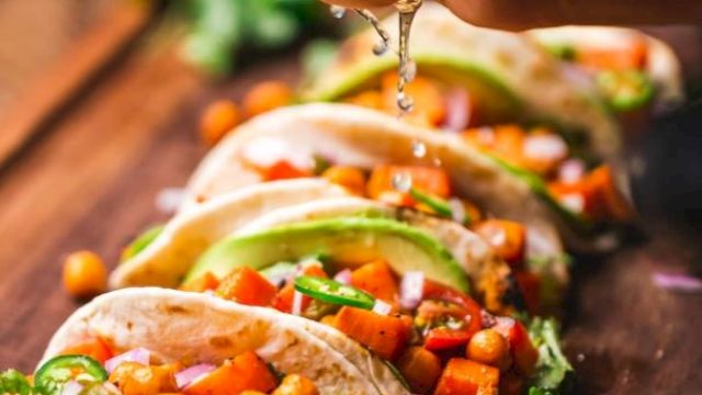 A person is squeezing lime over a row of chickpea and vegetable tacos on a wooden board, garnished with avocado and cilantro.
