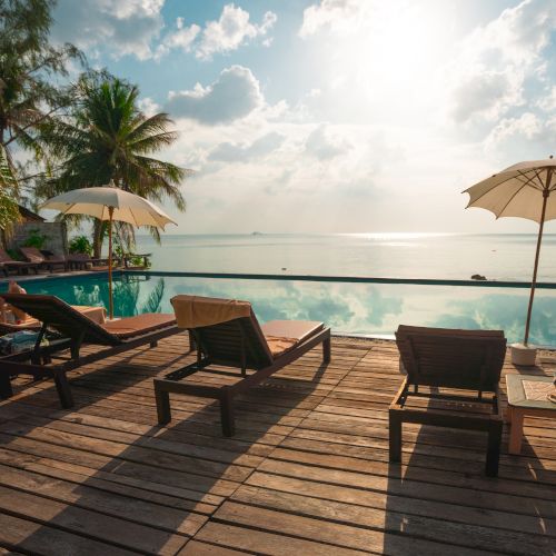 A tranquil poolside scene with lounge chairs, umbrellas, and palm trees overlooking the ocean under a bright sky, creating a serene atmosphere.