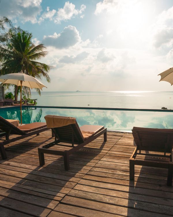 A tranquil poolside scene with lounge chairs, umbrellas, and palm trees overlooking the ocean under a bright sky, creating a serene atmosphere.