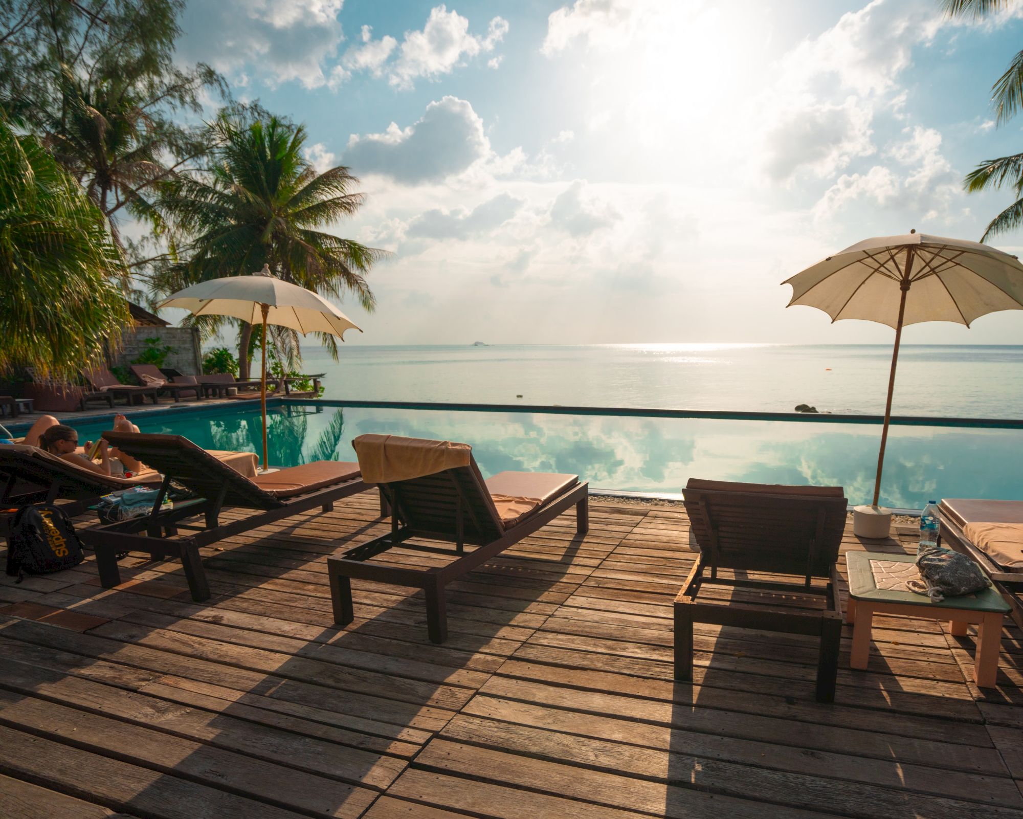 A serene beach scene with lounge chairs by a pool, umbrellas, palm trees, and a view of the ocean under a partly cloudy sky.