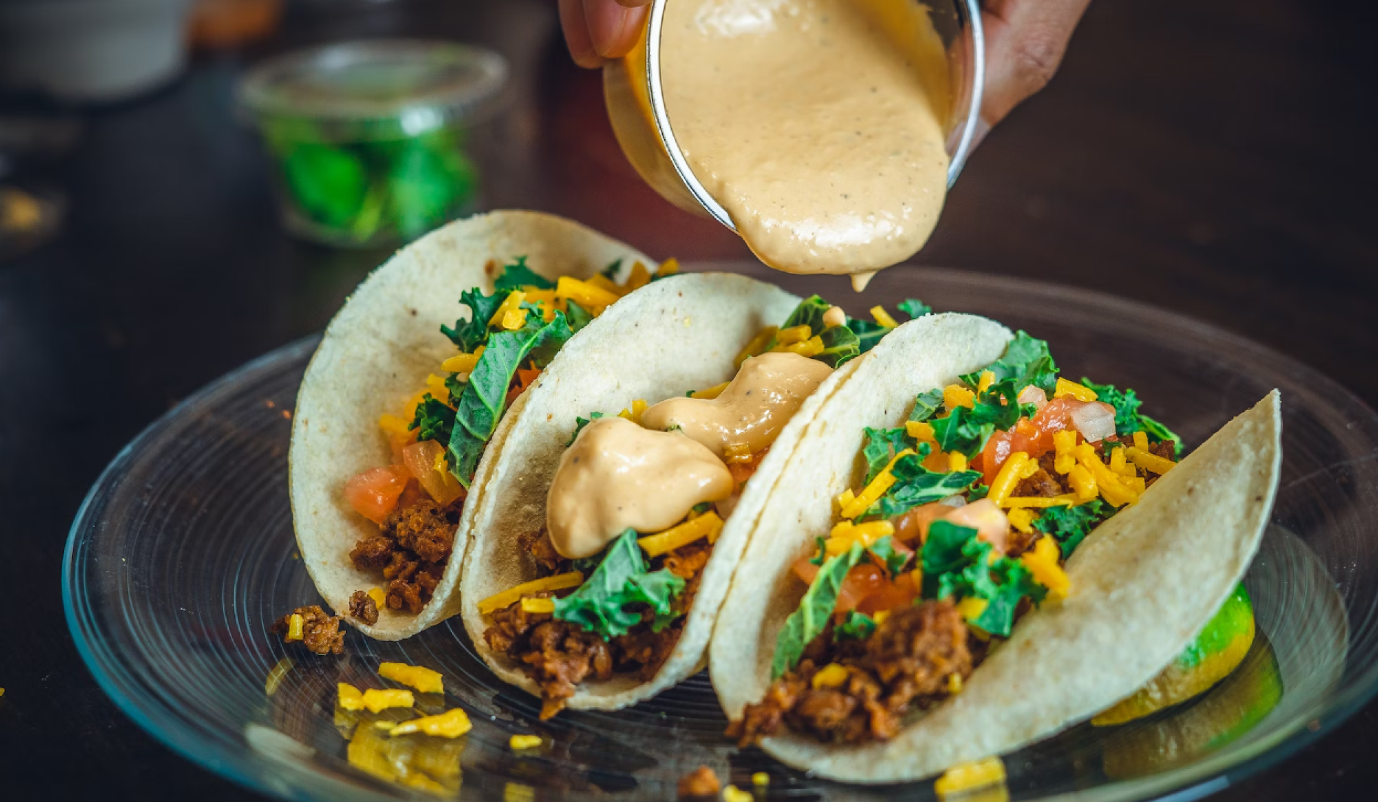 Three tacos with ground filling, cheese, kale, and tomatoes, drizzled with sauce, sit on a clear plate.