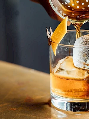 A cocktail being poured into a glass with ice and a garnish, on a bar counter.