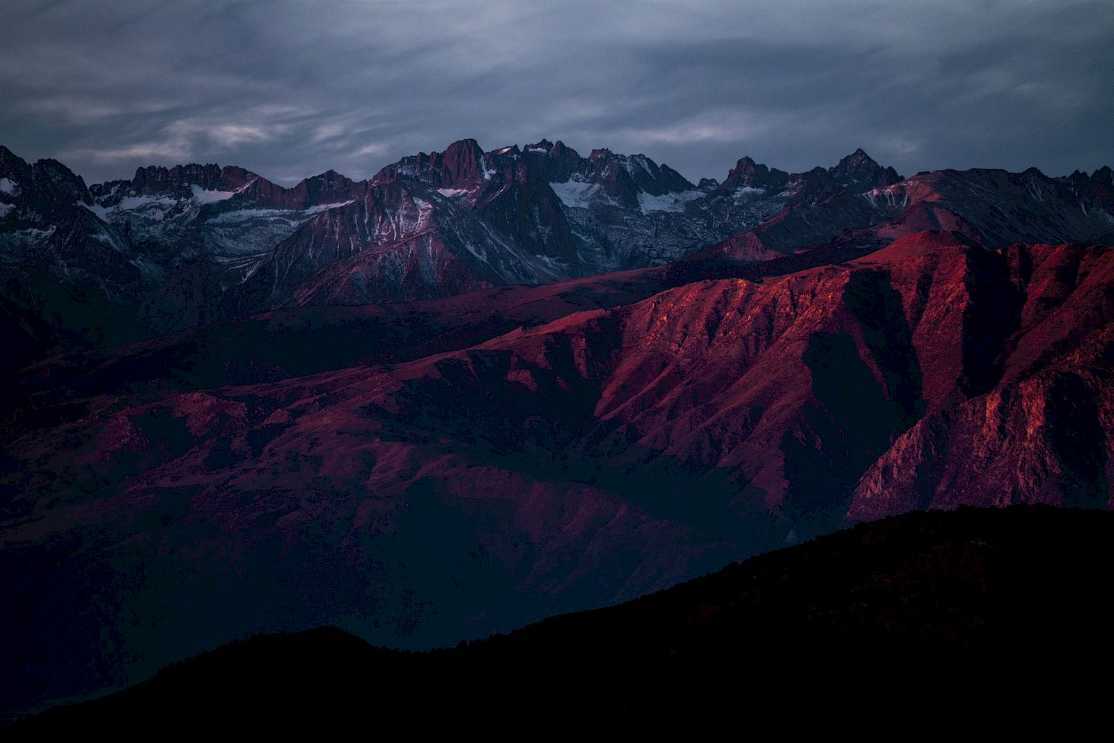 A mountain range at sunset with peaks partially covered in snow, bathed in a reddish hue under a cloudy sky.