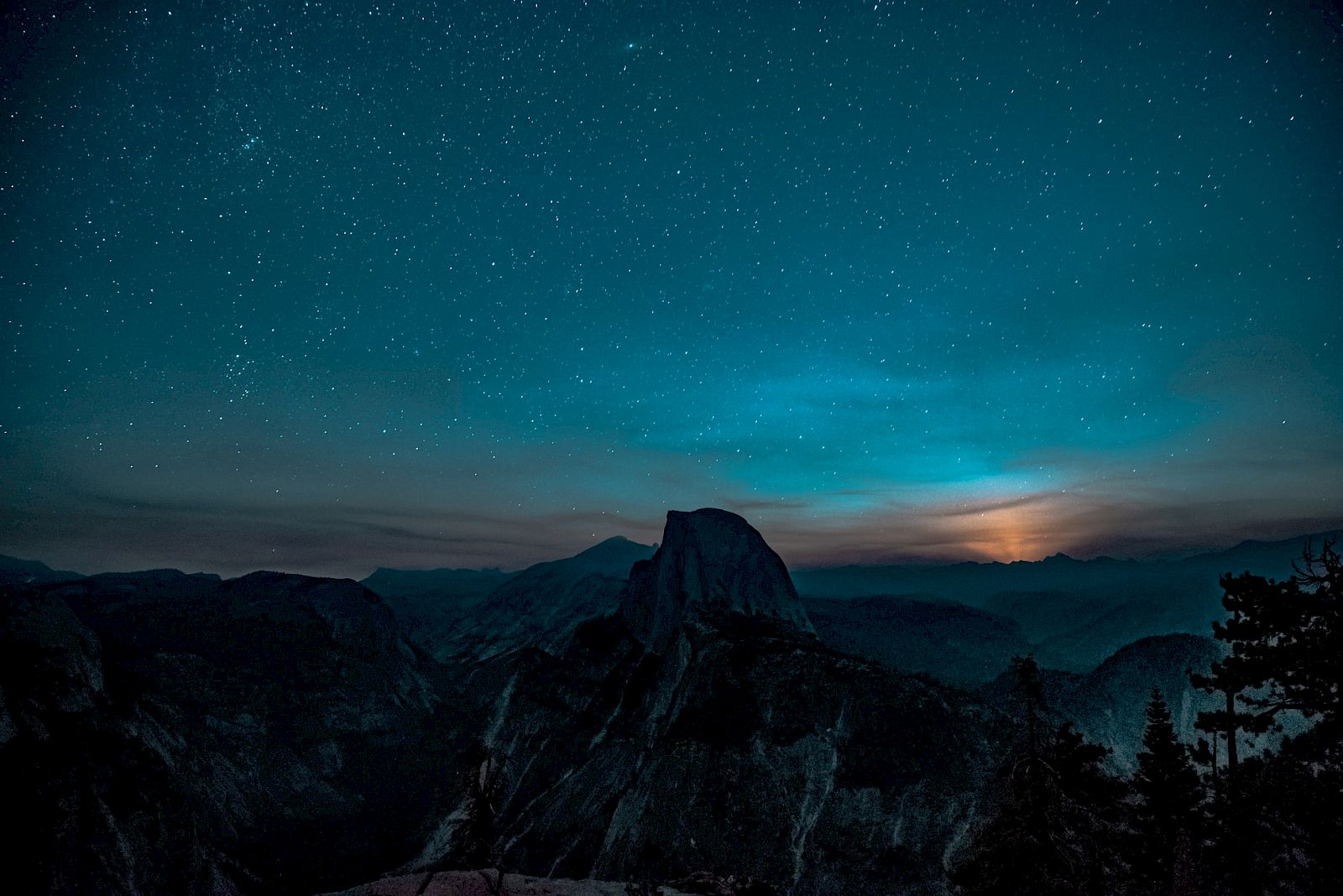 A starry night sky over a mountainous landscape with a glowing horizon and silhouetted trees and peaks in the foreground.