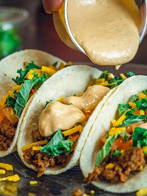 Three tacos with ground meat, cheese, lettuce, and diced tomatoes topped with sauce from a cup on a clear plate.