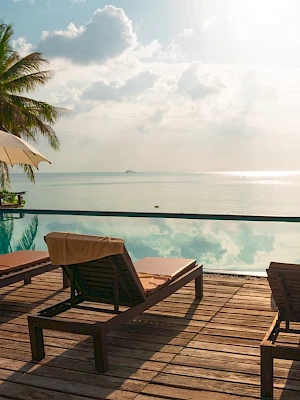 A tranquil poolside scene with lounge chairs, umbrellas, and a view of the ocean, surrounded by palm trees under a partly cloudy sky.