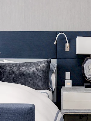 A neatly arranged bedroom with a blue and white theme, featuring a bed, side table, lamp, and wall-mounted reading light.