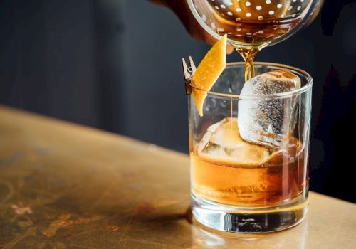A cocktail is being poured into a glass with ice and a citrus peel garnish, set on a wooden surface.