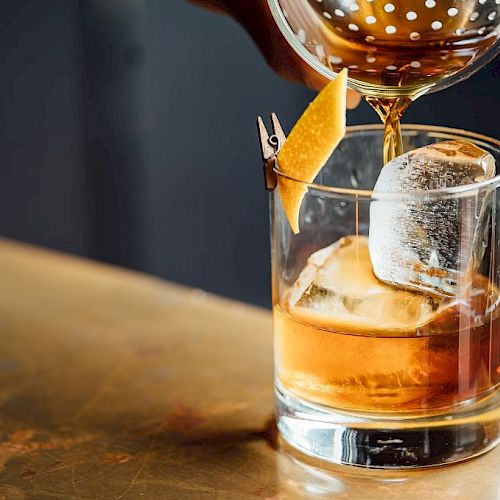 A cocktail is being poured into a glass with ice and a citrus peel garnish, set on a wooden surface.