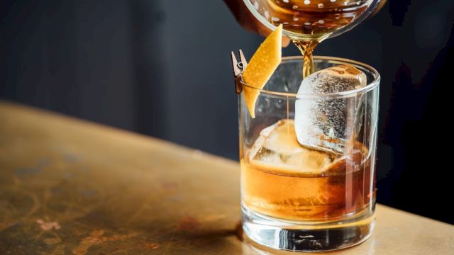 A cocktail is being poured into a glass with ice and a citrus peel garnish, set on a wooden surface.