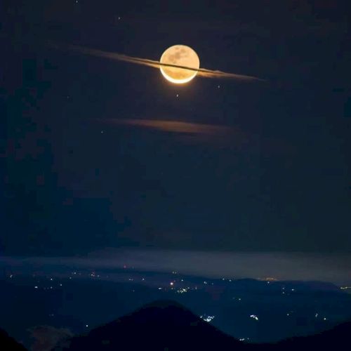 A full moon appears to have rings like Saturn due to cloud placement above a landscape with distant city lights at night.