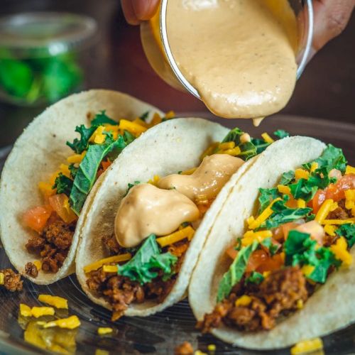 Three tacos with meat, cheese, lettuce, and sauce being poured on top, on a plate.