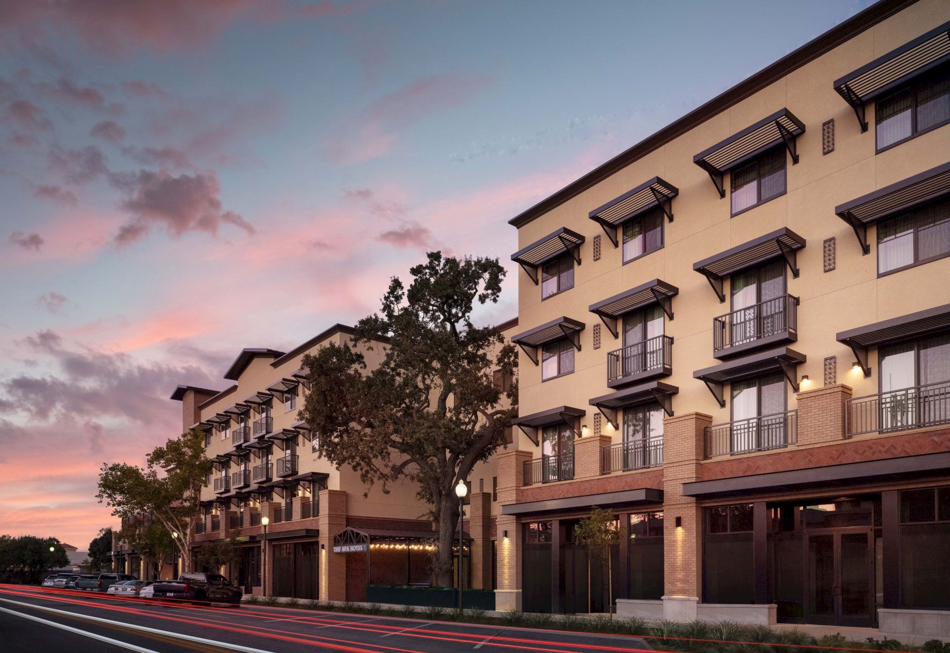 A modern multi-story hotel or apartment complex lined with balconies, trees, and a sunset sky, with a street and storefronts in front.
