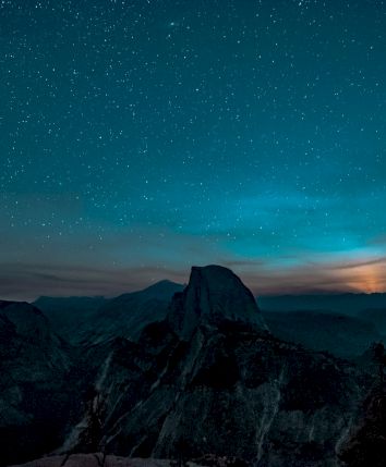 A starry night sky over a silhouette of mountains, with a glowing horizon suggesting a sunset or sunrise.