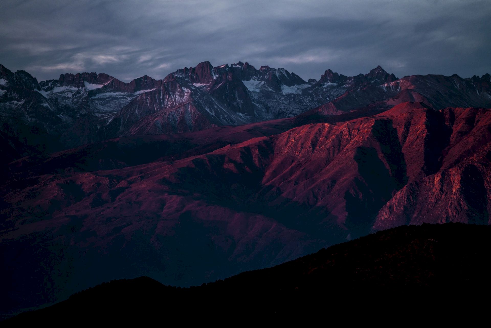 A mountain range illuminated by the warm glow of a sunset, with dramatic shadows and a cloudy sky.