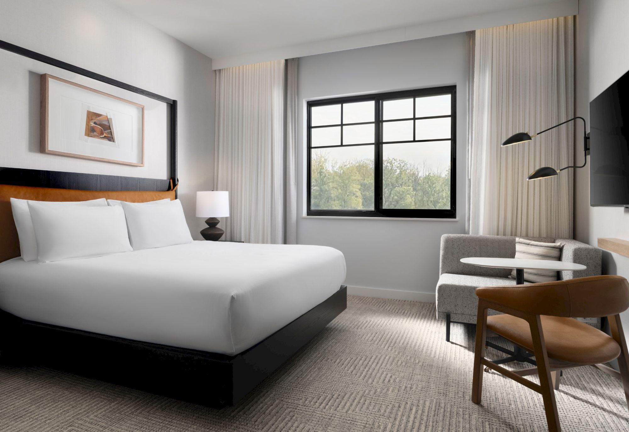 A modern hotel room with a large bed, sofa, table, chair, abstract art, and window with curtains. Neutral colors create a calm ambiance.