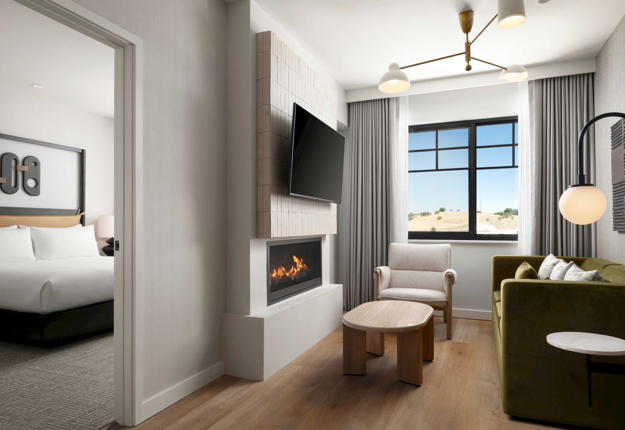 A modern living room with a fireplace, TV, and green sofa. A bedroom is visible through an open door.