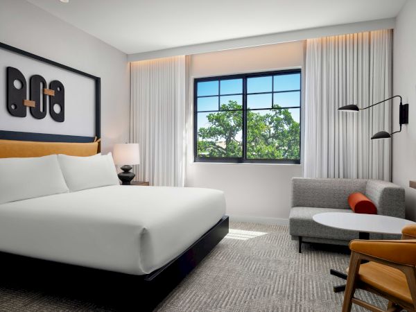 The image shows a modern hotel room with a bed, sofa, and window with greenery outside, decorated with abstract artwork and neutral tones.