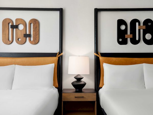 The image shows two beds in a hotel room with a lamp on a wooden nightstand between them and abstract wall art above each bed.
