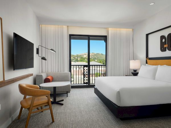 A modern hotel room with a bed, armchair, wall-mounted TV, and a balcony view overlooking a scenic landscape completes the decor.