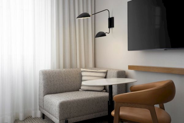 A modern room with a gray sofa, round table, brown chair, wall-mounted TV, and wall light fixture next to sheer curtains.