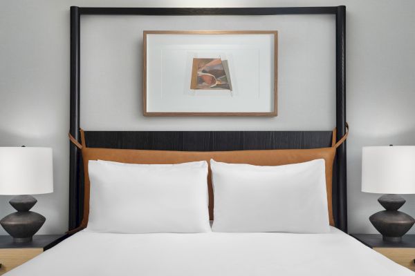 A neatly made bed with two white pillows, flanked by two lamps, and a framed abstract painting above the headboard.