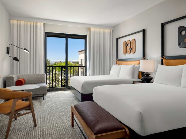 A hotel room features two beds, a modern sofa, a TV, art on the wall, and a balcony with a view outside.