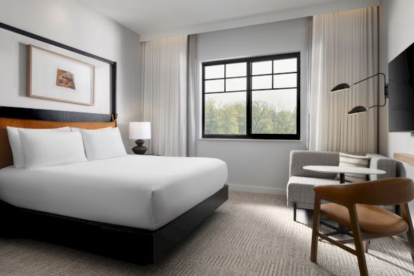 A modern hotel room with a large bed, a window, a lamp, a TV, and a seating area with a table and chairs in a cozy setup.