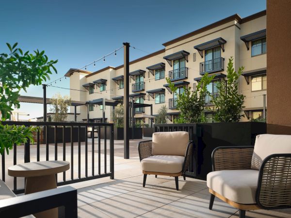 A sunny hotel rooftop patio with modern outdoor seating, potted trees, and string lights overlooking a courtyard.