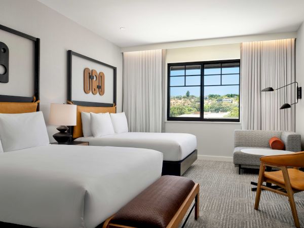A modern hotel room with two beds, a large window, art on the walls, and a seating area with a chair and side table.