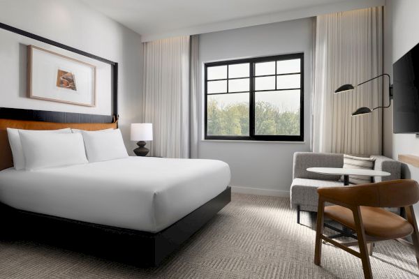 A modern hotel room features a bed, artwork, window, lamp, TV, and seating area with chairs and table, creating a cozy atmosphere.