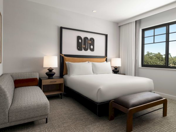 A modern bedroom with a large bed, lamps, artwork above the headboard, a window, a patterned carpet, and a gray sofa with a wooden bench.