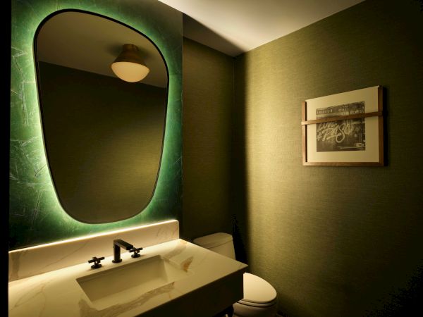 A modern bathroom with a sink, a backlit mirror, a wall-mounted picture, and a toilet on a dimly lit background.